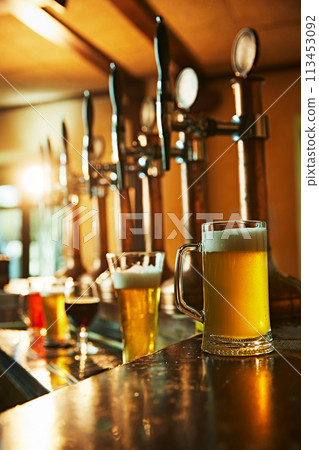 Shiny metal beer taps on wooden bar counter. Frothy mug of beer next to the taps with condensation on glass. Refreshment Shiny metal beer taps on wooden bar counter. Frothy mug of beer next to the taps with condensation on glass. Refreshment 113453092