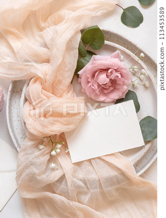 Card near cream fabric knot and light pink roses on plates top view copy space, wedding mockup 113453589