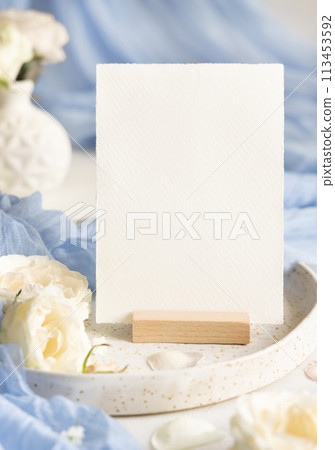 Card near light blue tulle fabric and cream flowers on plate close up, copy space, wedding mockup 113453592