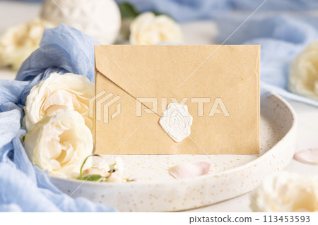 Sealed envelope near blue fabric and cream flowers on plate close up, copy space, wedding mockup 113453593