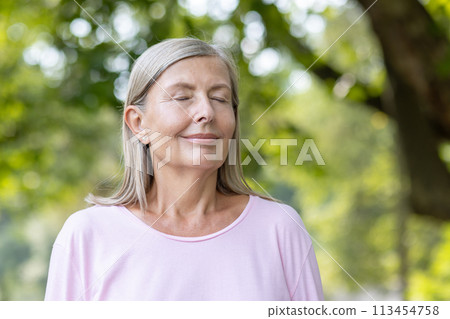 Close-up photo of an older woman standing in nature with her eyes closed and meditating and resting thoughtfully and relaxed. 113454758