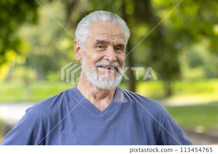 Close-up portrait of an older gray-haired man standing in a park in sportswear and smiling at the camera. 113454765