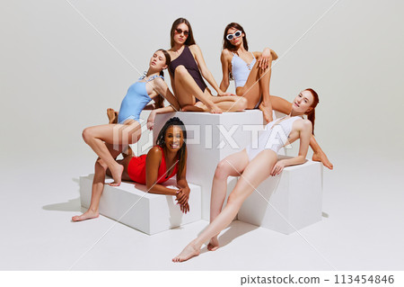 Fashion models in colorful swimsuits exhibit relaxed sophistication on minimalist cubes against white studio background. 113454846