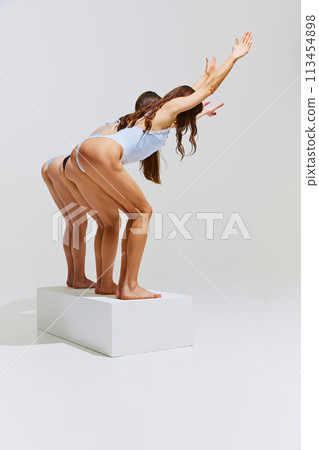 Back view of women in sleek swimsuits stands in diving posture on white platform against white studio background. 113454898