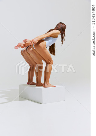 Rear view of women, friends in swimsuits simulate dive, blending sport with high fashion against white studio background. Rear view of women, friends in swimsuits simulate dive, blending sport with high fashion against white studio background. 113454904