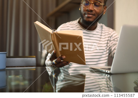 African American man holding notebook and using laptop at table in dinning room African American man holding notebook and using laptop at table in dinning room 113455587
