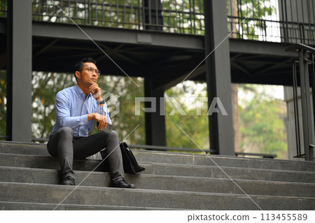 Full body of thoughtful adult businessman sitting on building steps and looking away Full body of thoughtful adult businessman sitting on building steps and looking away 113455589