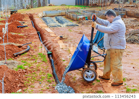 Using wheelbarrow, worker fills over drainage pipes with crushed stone Using wheelbarrow, worker fills over drainage pipes with crushed stone 113456243