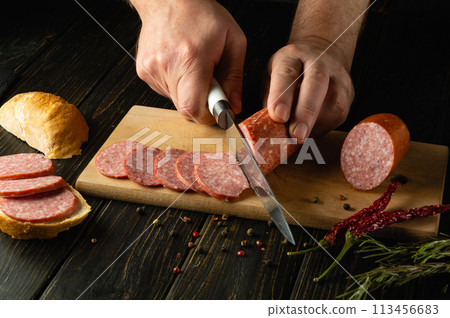 A man cuts veal sausage on a wooden cutting board. Preparing delicious sandwiches for dinner on the kitchen table at home A man cuts veal sausage on a wooden cutting board. Preparing delicious sandwiches for dinner on the kitchen table at home 113456683