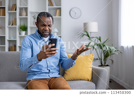 Perplexed African American man sitting in a living room, holding a smartphone and gesturing in confusion over a deceptive message. Perplexed African American man sitting in a living room, holding a smartphone and gesturing in confusion over a deceptive message. 113458300