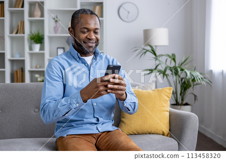 Relaxed young man browsing the internet on smartphone in a cozy living room setting, comfortable at home. 113458320