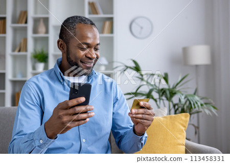 Casual man using smartphone and credit card for online shopping while sitting comfortably at home with indoor plants and bright interior. 113458331