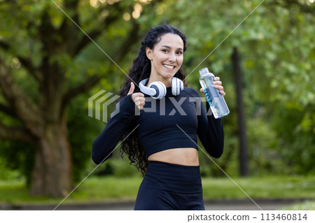 A cheerful Hispanic woman enjoys a fitness break at the park, holding a water bottle and wearing headphones, giving a thumbs up with trees in the background. A cheerful Hispanic woman enjoys a fitness break at the park, holding a water bottle and wearing headphones, giving a thumbs up with trees in the background. 113460814