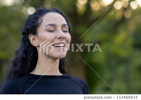 A hispanic woman closes her eyes with a content smile, basking in serenity amidst a natural green backdrop. A hispanic woman closes her eyes with a content smile, basking in serenity amidst a natural green backdrop. 113460833