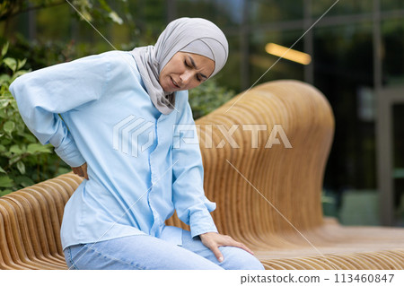 Diverse female person suffering from pain in back or reins due to incorrect posture while spending day outdoors. Unhappy woman feeling sudden acute ache in spinal muscles and leaning forward. Diverse female person suffering from pain in back or reins due to incorrect posture while spending day outdoors. Unhappy woman feeling sudden acute ache in spinal muscles and leaning forward. 113460847
