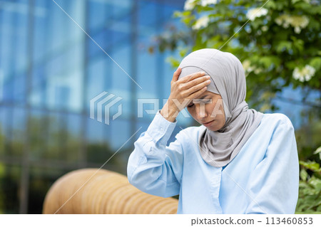 Sick woman in grey headscarf placing hand on forehead while sitting with closed eyes in bad condition. Stressed lady suffering from headache and sunstroke during unexpected heat in springtime. Sick woman in grey headscarf placing hand on forehead while sitting with closed eyes in bad condition. Stressed lady suffering from headache and sunstroke during unexpected heat in springtime. 113460853