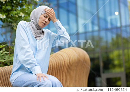 Stressed islamic woman having seat on bench while struggling with severe migraine outside. Exhausted female in cotton shirt grabbing head while feeling tensed and tired of high blood pressure. Stressed islamic woman having seat on bench while struggling with severe migraine outside. Exhausted female in cotton shirt grabbing head while feeling tensed and tired of high blood pressure. 113460854