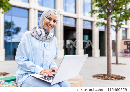 A cheerful Muslim woman wearing a hijab sits outdoors with a laptop, displaying productivity and connectivity in an urban setting. She's working near modern office buildings and trees. A cheerful Muslim woman wearing a hijab sits outdoors with a laptop, displaying productivity and connectivity in an urban setting. She's working near modern office buildings and trees. 113460859