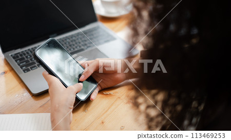 Close-up on the hands of a professional woman using a smartphone while working on a laptop at a wooden desk. 113469253