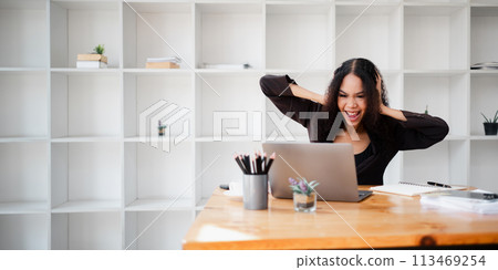 Excited businesswoman with arms raised in a victory gesture, celebrating a successful moment while working on her laptop in the office. 113469254