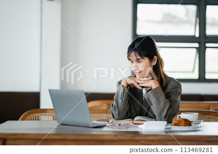 Contemplative young woman gazes at her laptop screen, deep in thought, in a cozy workplace with a coffee and croissant on the table. 113469281