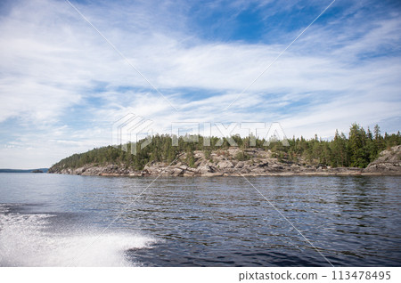 Rocky and stone river bank with fir trees on a sunny day. Water splashes from the boat, calm 113478495