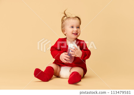 Little baby girl, child with ponytails in red dress and socks holding bottle, smiling, sitting on beige background. 113479581