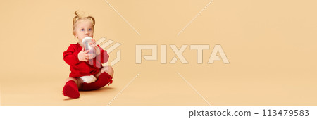 Little baby girl, child with ponytails in red dress and socks holding bottle, smiling, sitting on beige background. Little baby girl, child with ponytails in red dress and socks holding bottle, smiling, sitting on beige background. 113479583