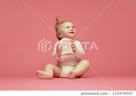 Smiling baby in comfortable onesie sitting on pink background, holding moisturizing skin cream. Cosmetics for babies 113479587