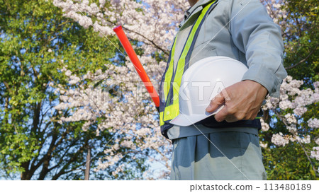 A man in work clothes holding a guide wand and a helmet with spring cherry blossoms in the background | Image of construction and security work A man in work clothes holding a guide wand and a helmet with spring cherry blossoms in the background | Image of construction and security work 113480189
