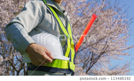 A man in work clothes holding a guide wand and a helmet with spring cherry blossoms in the background | Image of construction and security work 113480192