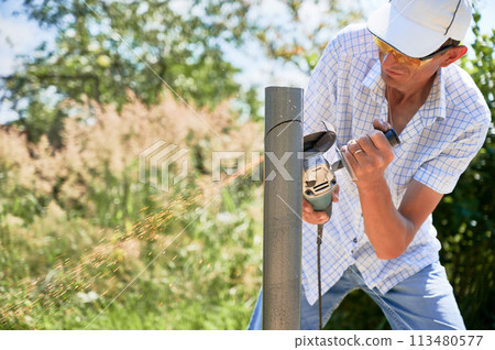 Male worker cutting metal pole with electric grinder. Man in protective glasses using hand-held grinder while preparing aluminum pole for solar panel installation. 113480577