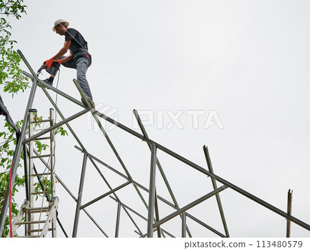 Man engineer standing on metal rail and using electric drill while installing support structure for photovoltaic solar panels. Isolated on white background. Man engineer standing on metal rail and using electric drill while installing support structure for photovoltaic solar panels. Isolated on white background. 113480579