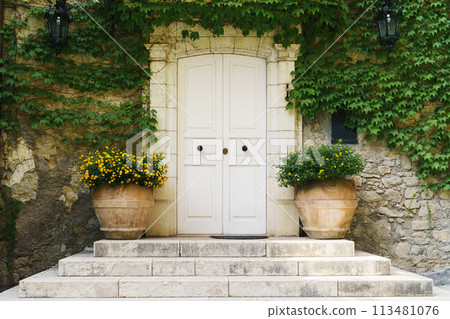 Beautiful white doors with decorative green plants and flowers near the house's stone walls. 113481076