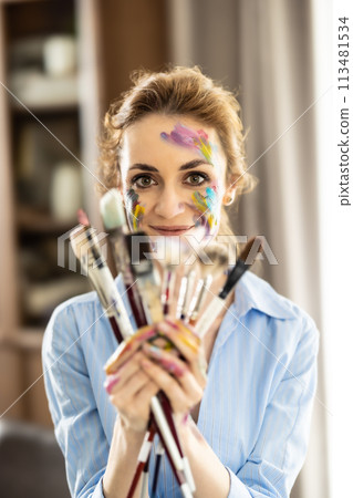 Portrait of a cute Latin woman holding a bunch of paintbrushes with both hands and smiling 113481534