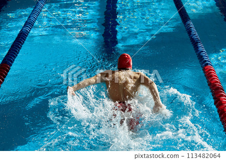 Top view of muscular, athletic young man, swimmer in red cap in motion, showing strength, training, swimming in pool indoors 113482064