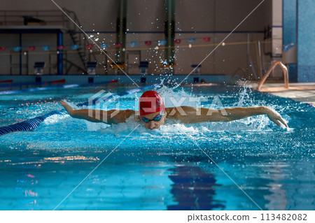 Young man in red cap and goggles, swimmer in motion, showing butterfly stroke, training, swimming in pool indoors Young man in red cap and goggles, swimmer in motion, showing butterfly stroke, training, swimming in pool indoors 113482082