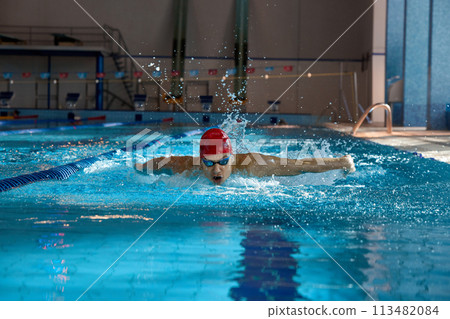 Young man in red cap and goggles, swimmer in motion, showing butterfly stroke, training, swimming in pool indoors Young man in red cap and goggles, swimmer in motion, showing butterfly stroke, training, swimming in pool indoors 113482084