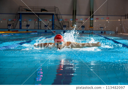 Competitive, muscular young man, swimmer in red cap performs butterfly stroke in pool, training indoors Competitive, muscular young man, swimmer in red cap performs butterfly stroke in pool, training indoors 113482097