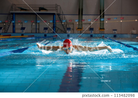 Competitive swimmer in red cap and goggles in motion, training butterfly stroke technique, swimming in pool Competitive swimmer in red cap and goggles in motion, training butterfly stroke technique, swimming in pool 113482098