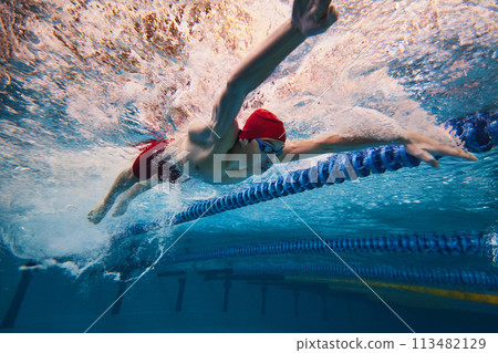 Strength and resilience. Young man, swimmer in red cap and goggles swimming, training in pool indoors. Strength and resilience. Young man, swimmer in red cap and goggles swimming, training in pool indoors. 113482129