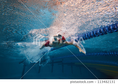 Strength and resilience. Young man, swimmer in red cap and goggles swimming, training in pool indoors. Strength and resilience. Young man, swimmer in red cap and goggles swimming, training in pool indoors. 113482131