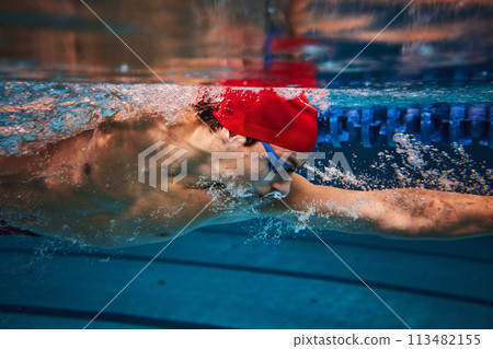 Concentration. Competitive young man, swimming athlete in goggles and red cap in motion, training in pool indoors. Concentration. Competitive young man, swimming athlete in goggles and red cap in motion, training in pool indoors. 113482155