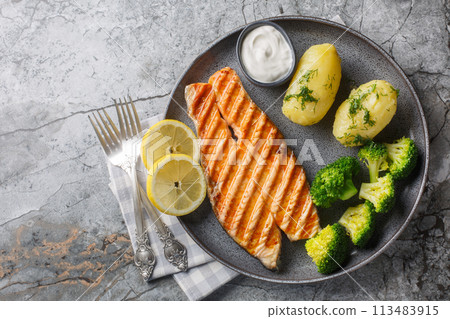 Dietary grilled salmon with boiled potatoes, broccoli, lemon, herbs and cream sauce close-up in a plate. Horizontal top view 113483915
