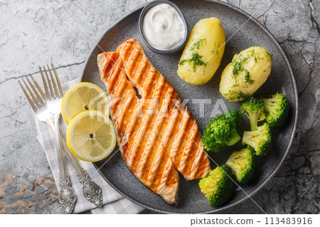 Delicious grilled salmon steak with boiled new potatoes, broccoli, lemon and cream sauce close-up in a plate. Horizontal top view Delicious grilled salmon steak with boiled new potatoes, broccoli, lemon and cream sauce close-up in a plate. Horizontal top view 113483916