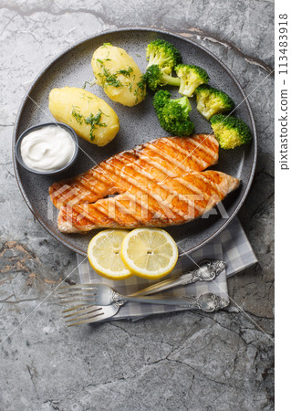 Close up of delicious grilled salmon with new potatoes, broccoli and cream sauce in a plate. Vertical top view Close up of delicious grilled salmon with new potatoes, broccoli and cream sauce in a plate. Vertical top view 113483918