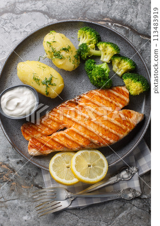 Grilled salmon steak with new potatoes, broccoli and cream sauce close-up in a plate. Vertical top view Grilled salmon steak with new potatoes, broccoli and cream sauce close-up in a plate. Vertical top view 113483919