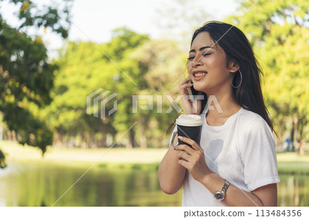 Asian Woman use smartphone drinking coffee hand holding hot disposable cup in green park. Banner Happy Relax asian woman smiling face at outdoors garden. Young women enjoy nature with copy space Asian Woman use smartphone drinking coffee hand holding hot disposable cup in green park. Banner Happy Relax asian woman smiling face at outdoors garden. Young women enjoy nature with copy space 113484356