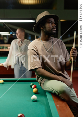 Side view portrait of Black adult man wearing hat posing on billiards table in bar 113485199