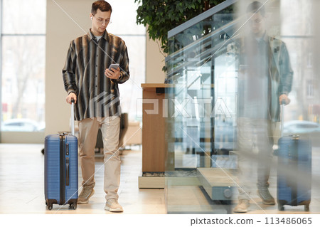 Full length portrait of modern young man with suitcase walking towards camera in hotel lobby and using smartphone with taxi app copy space 113486065